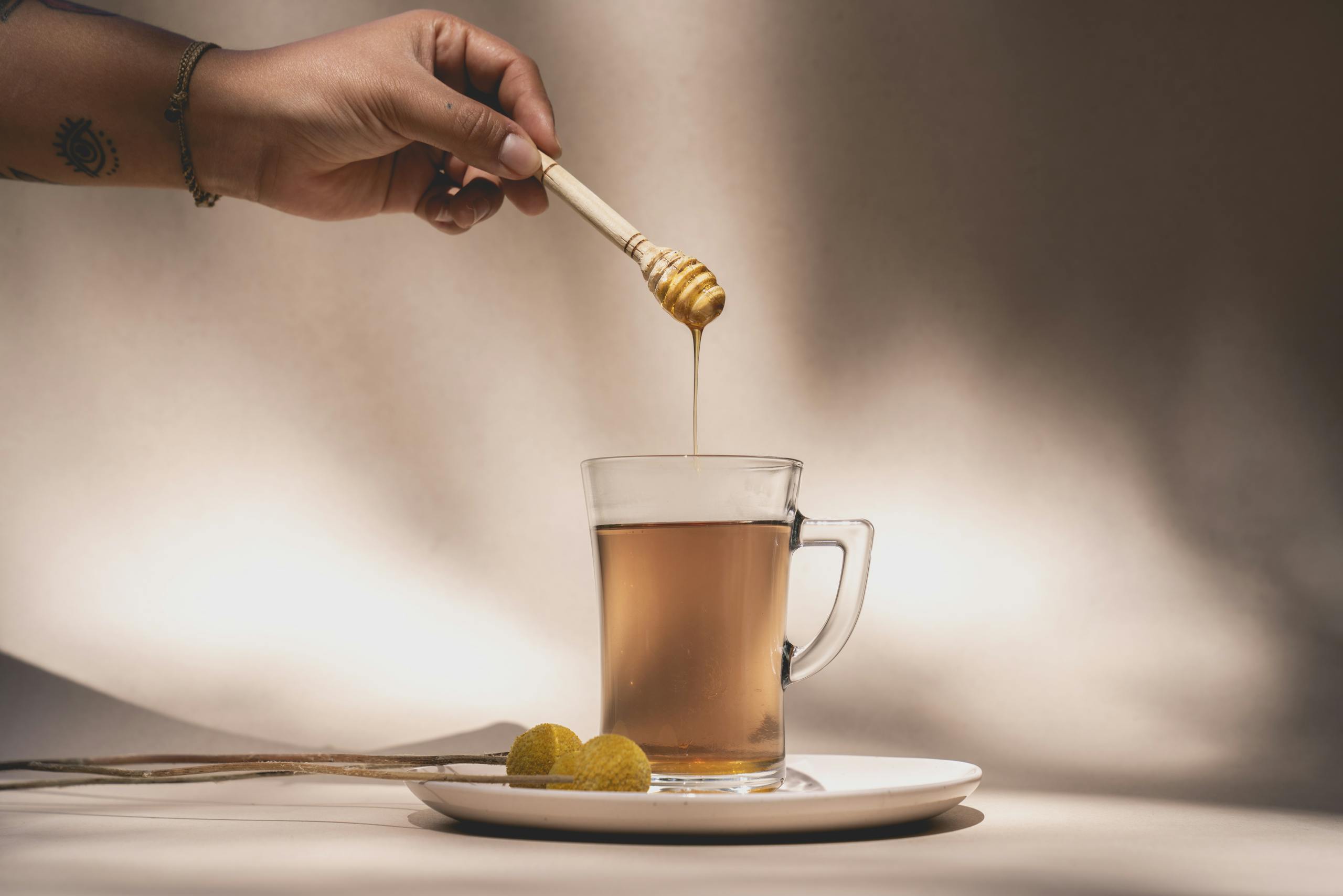 A hand drips honey into a glass mug of tea using a honey dipper, with a warm, inviting atmosphere.